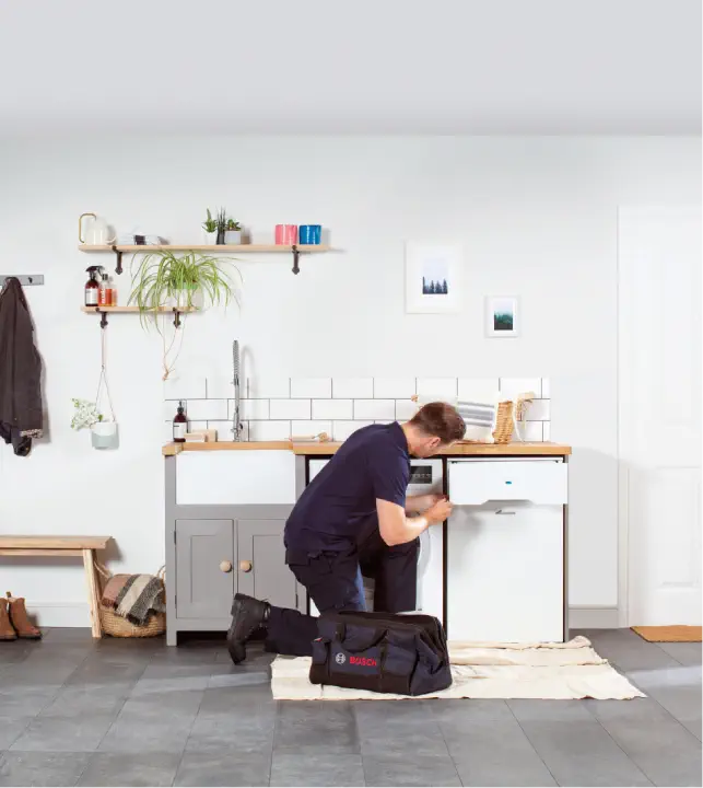 A technician repairs an oil boiler under a sink in a modern kitchen. Tools are spread on a cloth, with a tool bag labeled "Bosch" beside him. The kitchen, equipped with advanced home heating systems, has minimal decor and plants on a shelf.